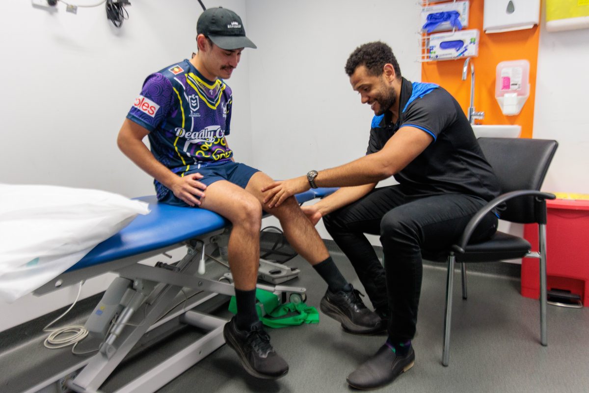 A client wearing a Deadly Choices shirt and black cap sits on an examination table while our deadly physiotherapist Joshua Weier, dressed in black with blue accents, examines the client’s knee. They are in a physiotherapy room with various medical supplies and equipment visible in the background.