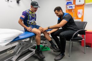 A client wearing a Deadly Choices shirt and black cap sits on an examination table while our deadly physiotherapist Joshua Weier, dressed in black with blue accents, examines the client’s knee. They are in a physiotherapy room with various medical supplies and equipment visible in the background.