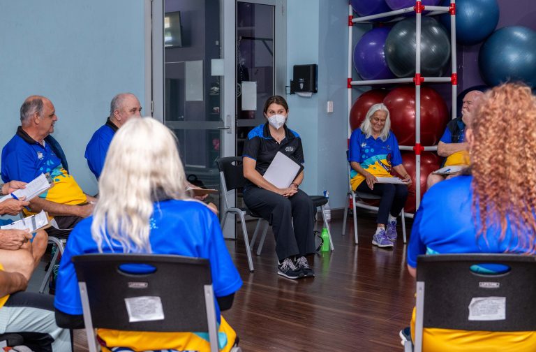 A Work It Out facilitator leads a group session with Elders seated in a circle, discussing health and exercise goals in a supportive indoor setting.
