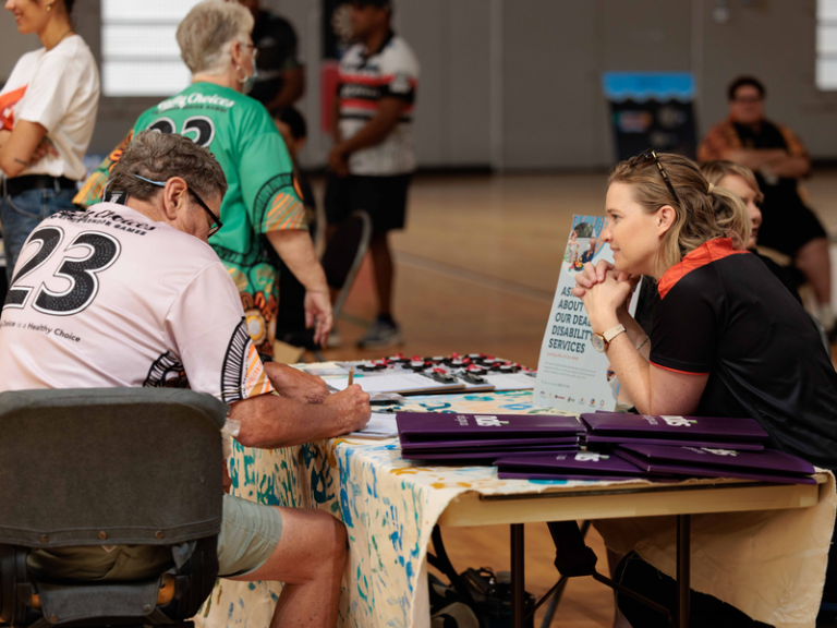 An IUIH Allied Health team member sits at a table speaking with a Community member during a disability services stall, with Deadly Choices shirts and information packs visible.