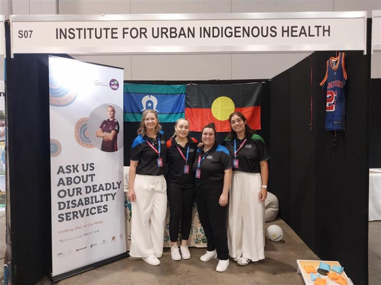 Four IUIH Support Coordinators standing together at a community event booth, smiling in front of Aboriginal and Torres Strait Islander flags and a display promoting Deadly Disability Services.