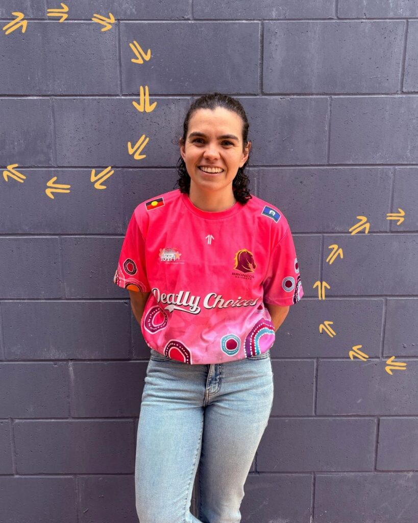 Kara O'Sullivan, an OT at the Institute for Urban Indigenous Health, stands against a grey wall. She is smiling.